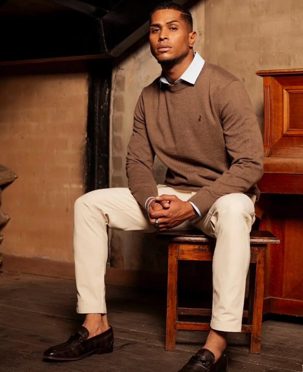 A New Era Africa, Founder and Executive Director. Lyle Volkwyn. A man sitting on a wooden chair in a rustic room, dressed in a brown sweater, white pants, and dark loafers, with a serious expression.