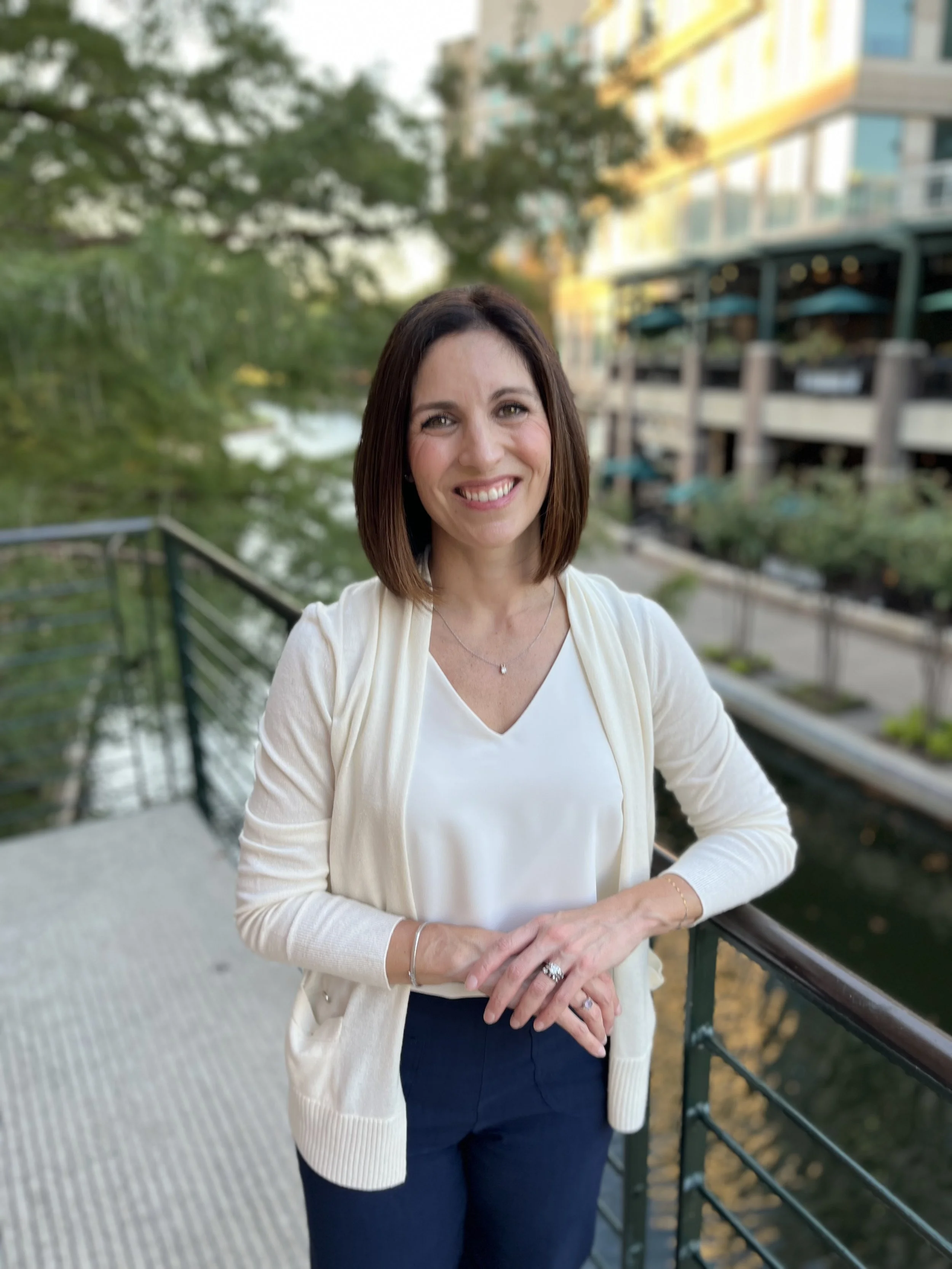 Heather Gallagher, a smiling woman with shoulder-length brown hair standing outdoors on a balcony with a drink in her hand, wearing a white blouse and cream cardigan, with trees and a modern building in the background.