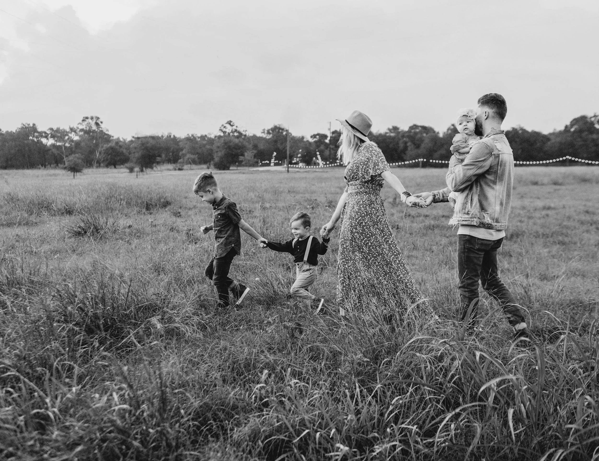 A black and white photo of a family walking through a grassy field holding hands, with a woman in a hat, two children, and two men carrying a young girl.