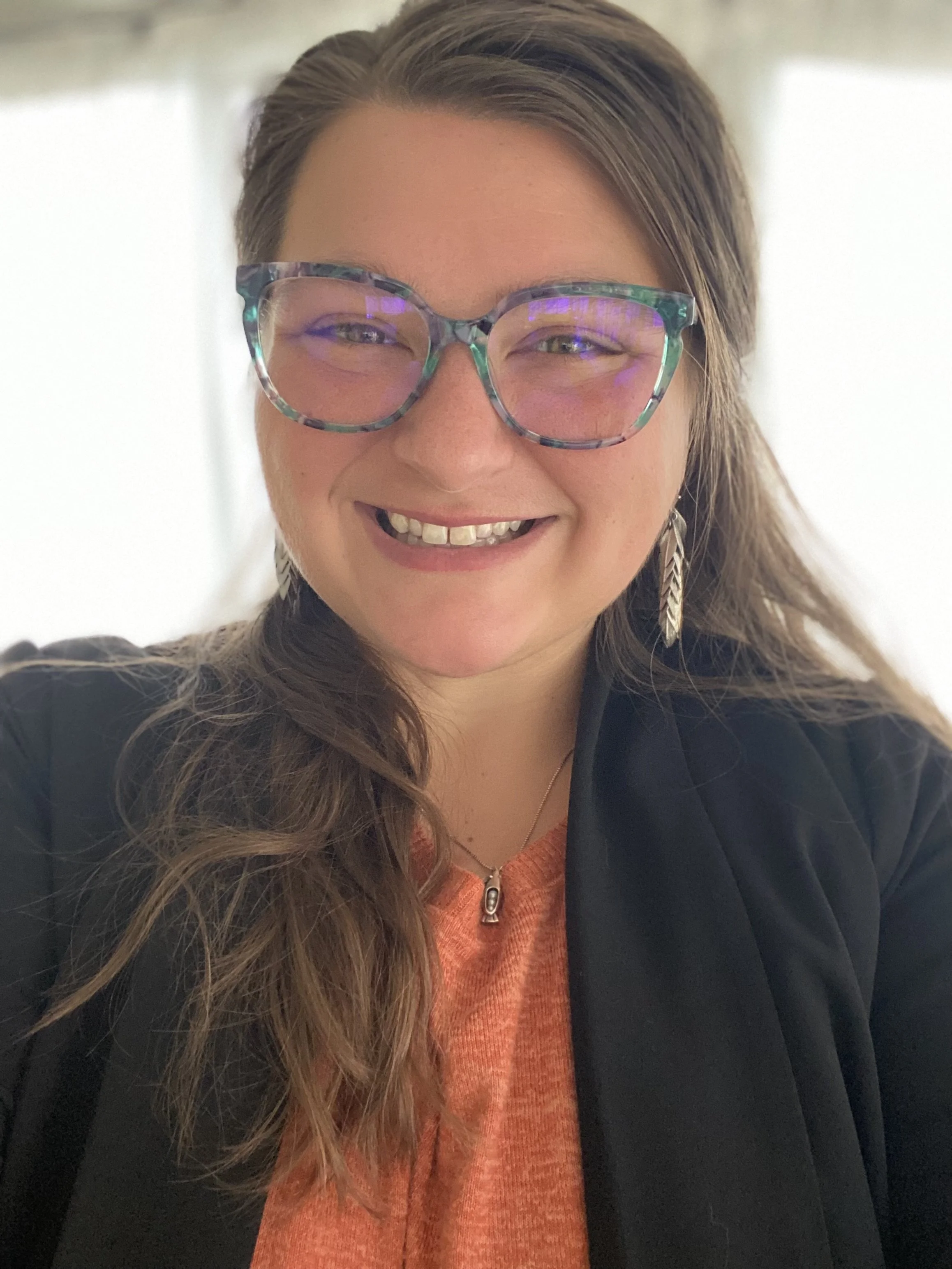 Close-up of a smiling woman with long brown hair, wearing colorful glasses, a black jacket, an orange top, and a feather earring.
