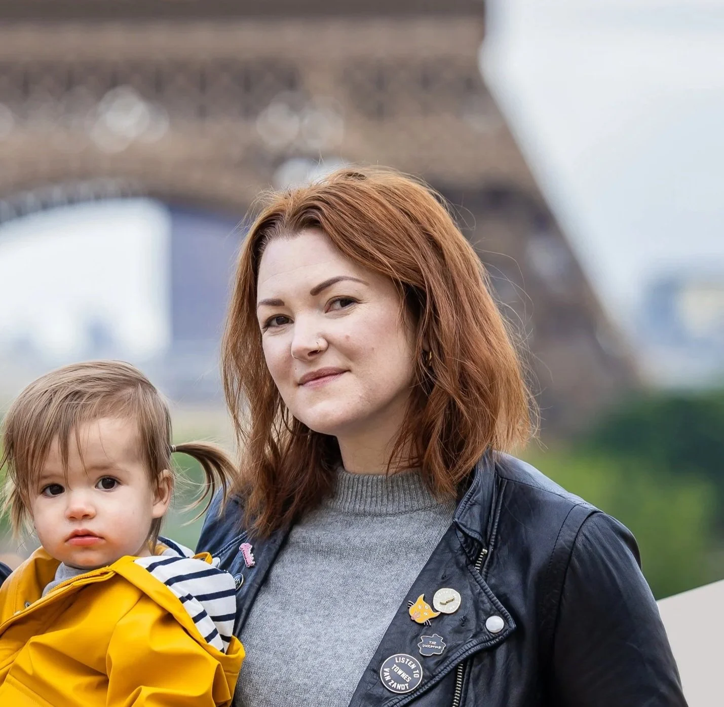 Photo of woman with auburn hair from the shoulders up wearing a black leather jacket and colorful pins