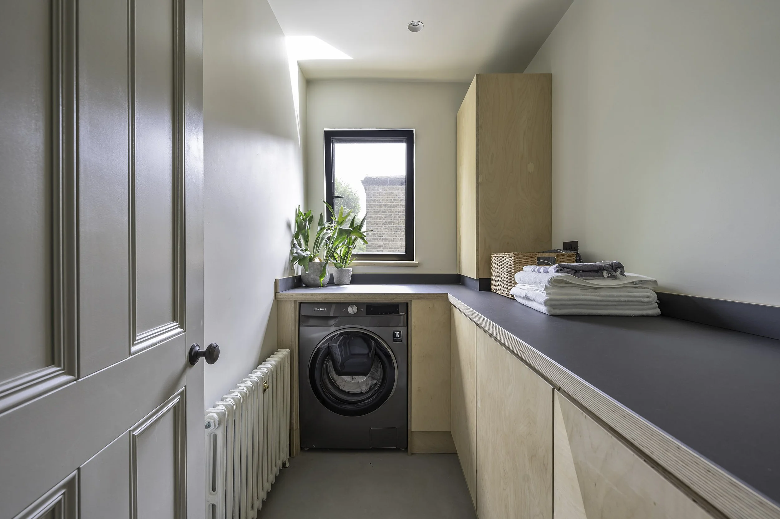 Bespoke Laundry utility room with in birch plywood, with a front-loading washing machine, a black countertop with folded towels, a window with plants on the sill, and light wood cabinetry.