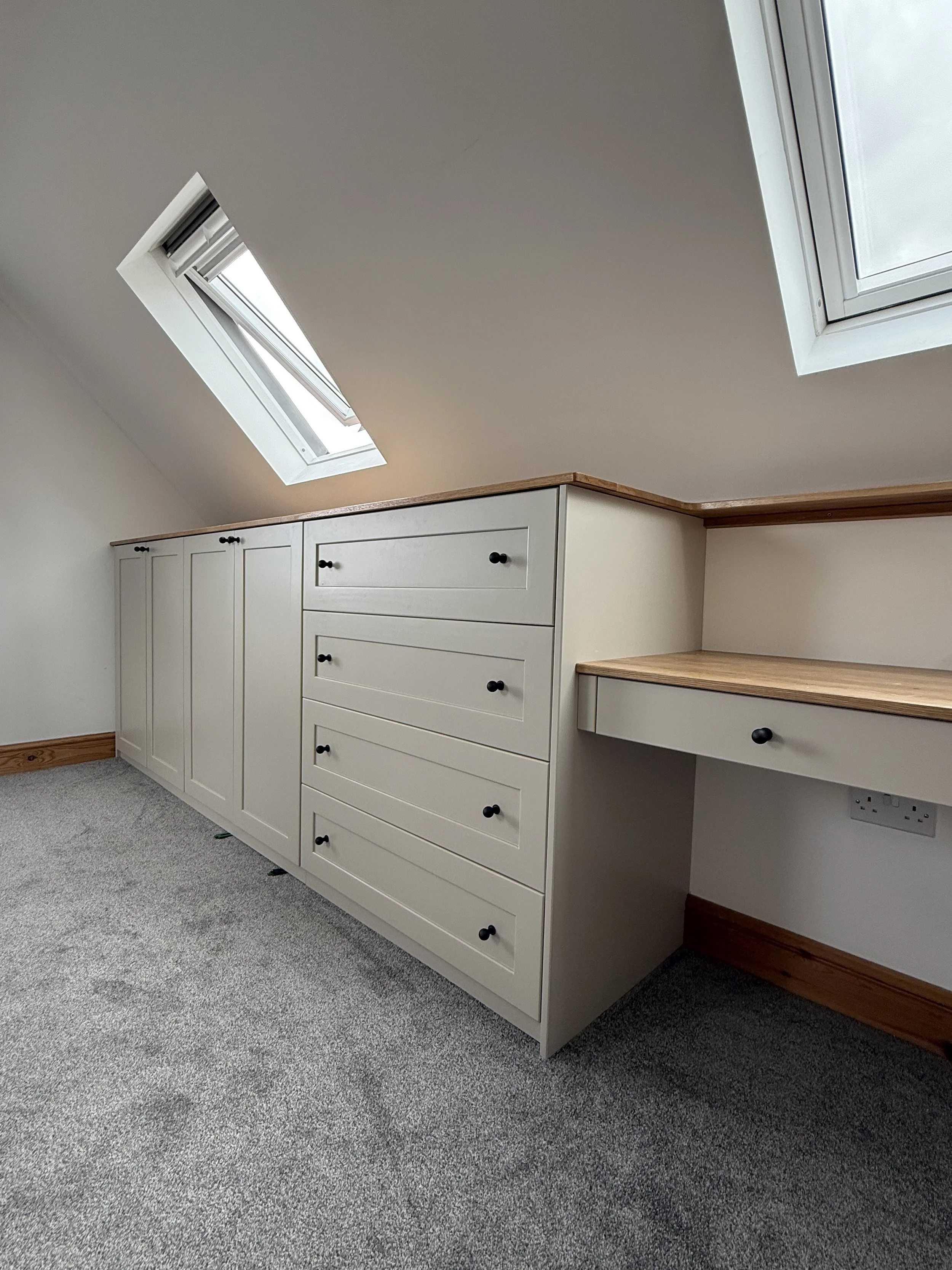 A built-in bespoke attic white cabinet with bronze Corston Architectural knobs underneath two skylight windows on a sloped ceiling. The cabinet has drawers and doors, with a wooden top surface extending to the right side.