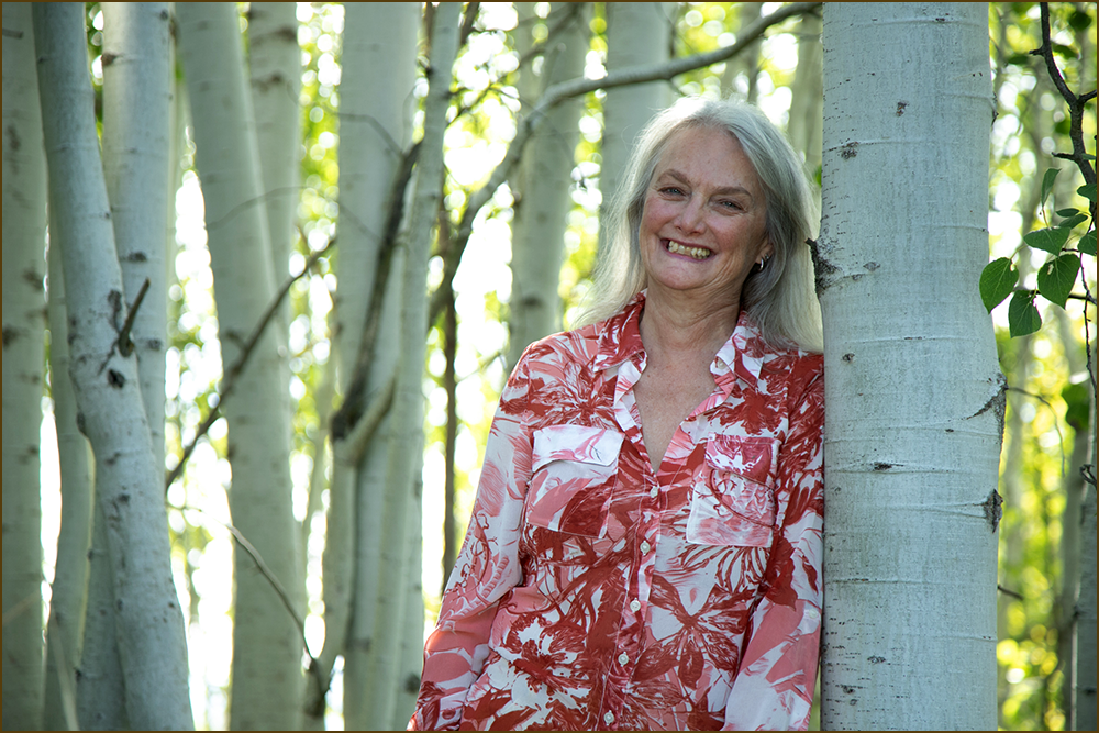 Louise Sidley, author - with a cheerful expression, wearing a red and white patterned shirt, standing outdoors amidst tall aspen trees with bright sunlight filtering through the leaves.