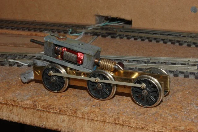 A small model train locomotive with a motor and wheels on a wooden surface, positioned near train tracks.