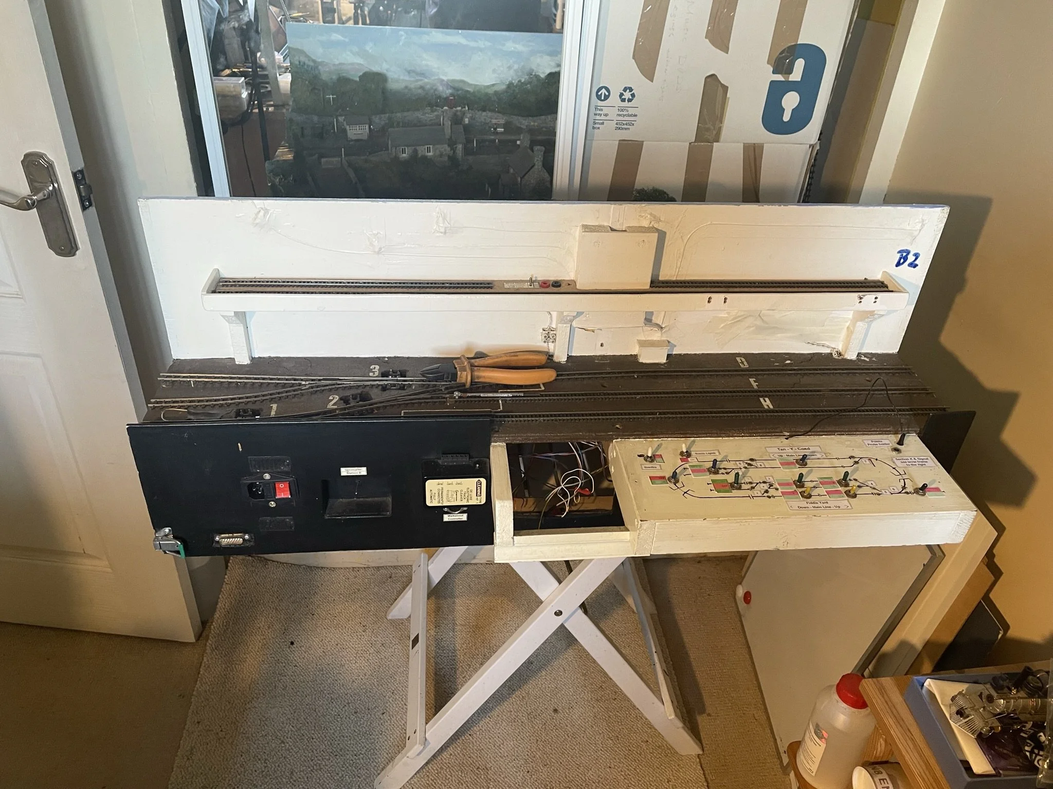 Model railway track setup with electrical components and wiring, placed on a white table in front of a glass door and cardboard boxes.