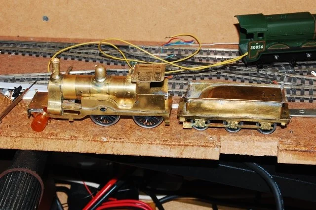 Brass model steam train with an engine and tender on a wooden track, connected to a green locomotive at the back.