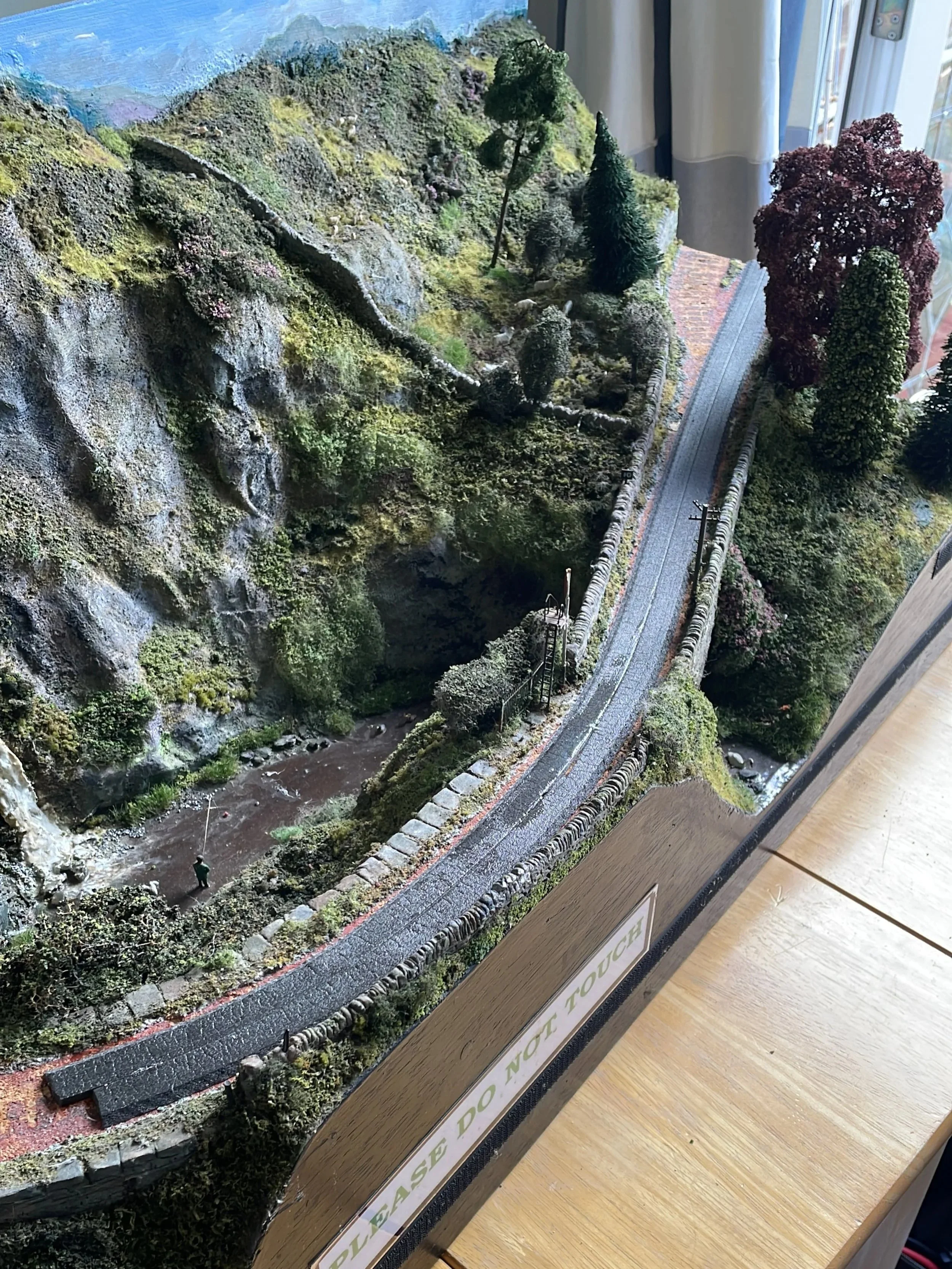 Detailed miniature landscape model depicting a rocky hillside, trees, a small river, and a bridge over a rail track with a sign that says 'Please do not touch'.