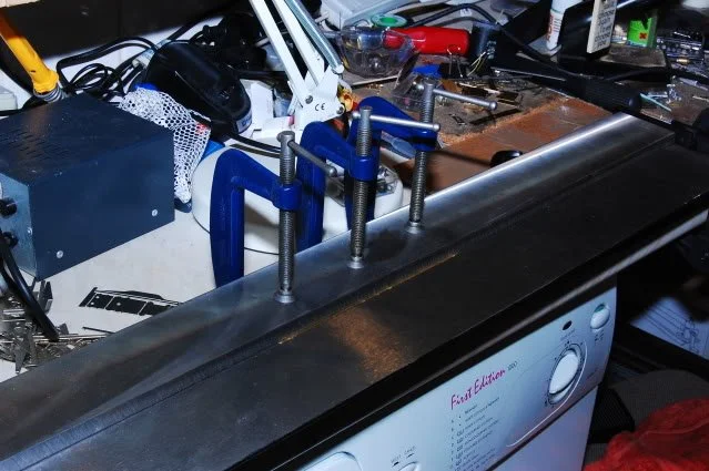 A close-up of a metal work surface with blue clamps holding a metal piece, surrounded by tools, wires, and electronic components indicating a workspace for electronics or metalwork projects.