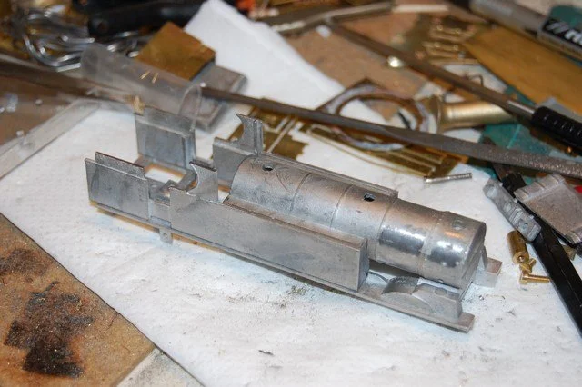 A metal model of a steam locomotive under construction, surrounded by tools and work materials.