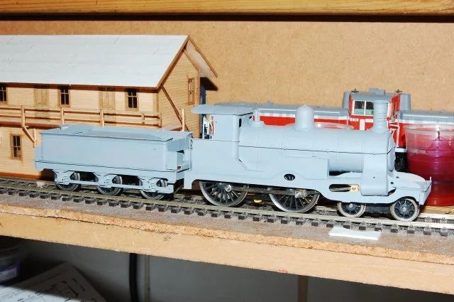 A blue model steam locomotive train on tracks, with a wooden building and a red container in the background.