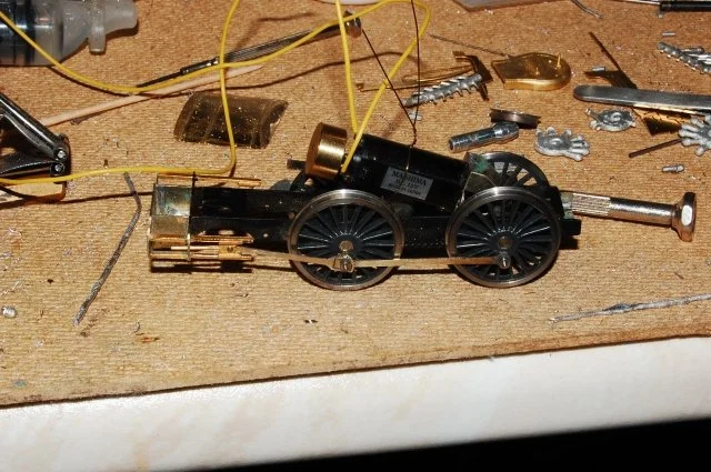 A small black and gold model train engine with yellow wires, surrounded by scattered small metal parts and tools on a workbench.