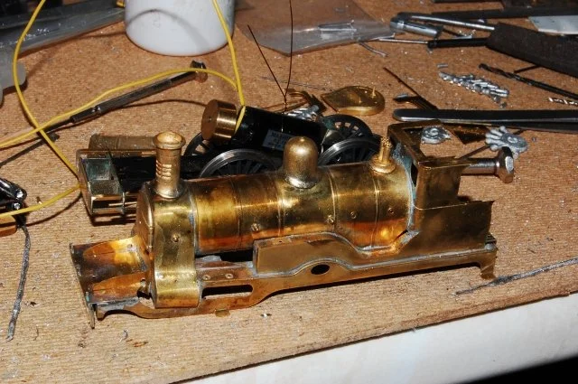 Close-up of a brass model steam engine on a workbench with wires, tools, and small parts around it.
