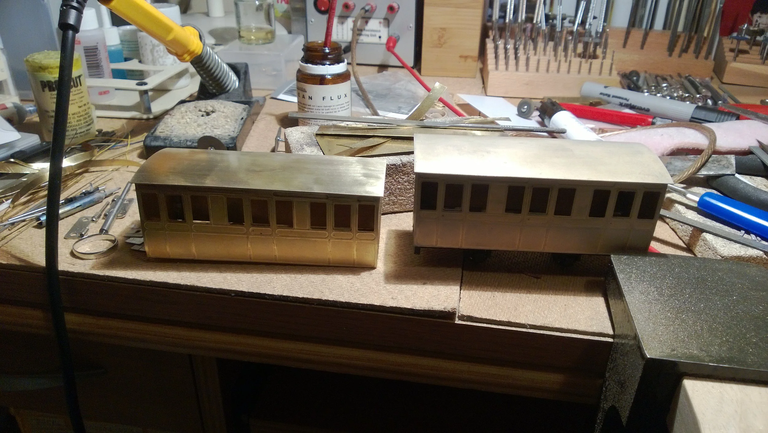 Two model train cars, one gold-colored with windows and one uncolored, on a workbench surrounded by tools and materials for craft or model building.