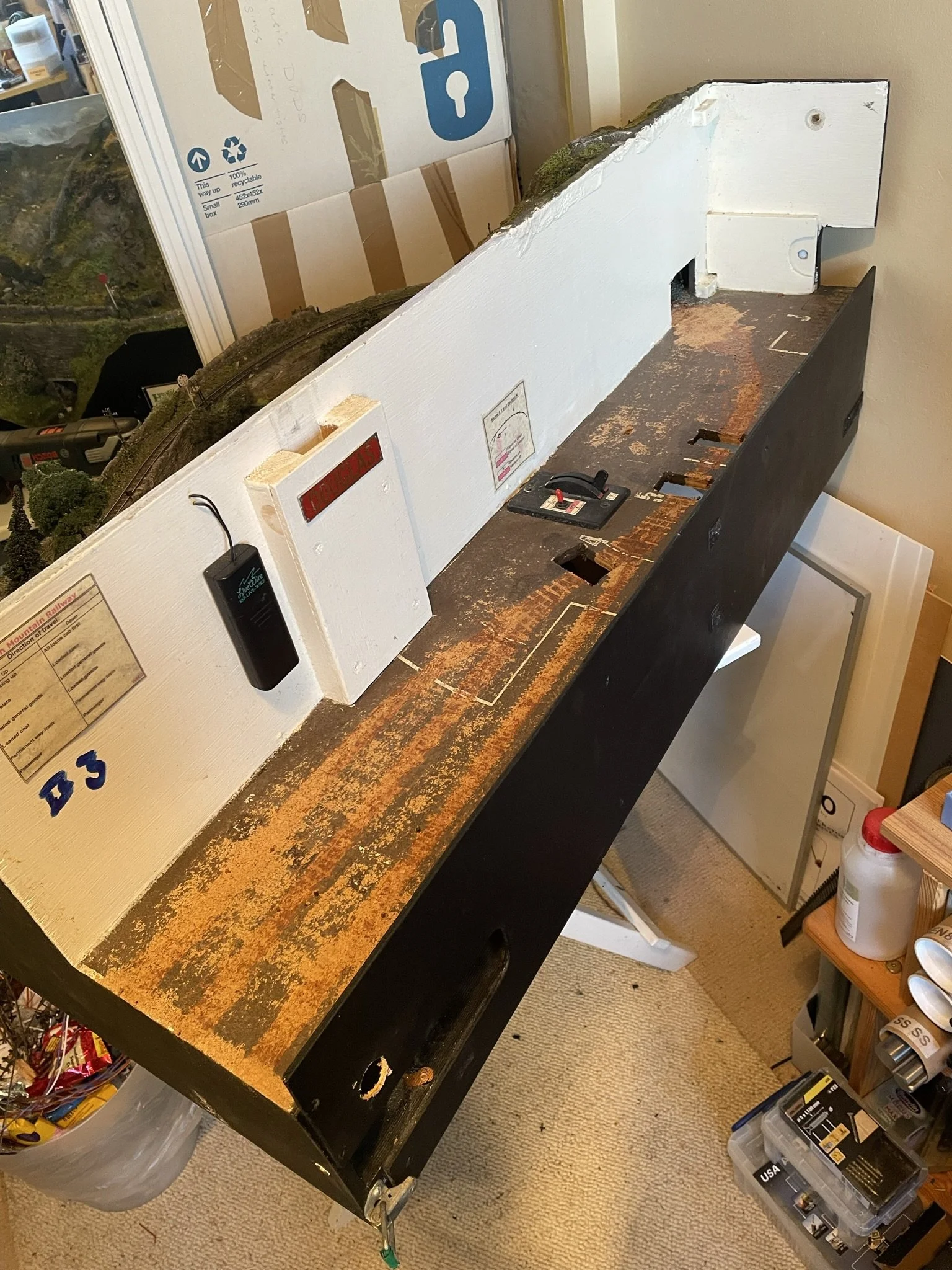 Model train setup on a black table with a white wooden backdrop, some track sections, and greenery. Various tools and supplies are nearby.