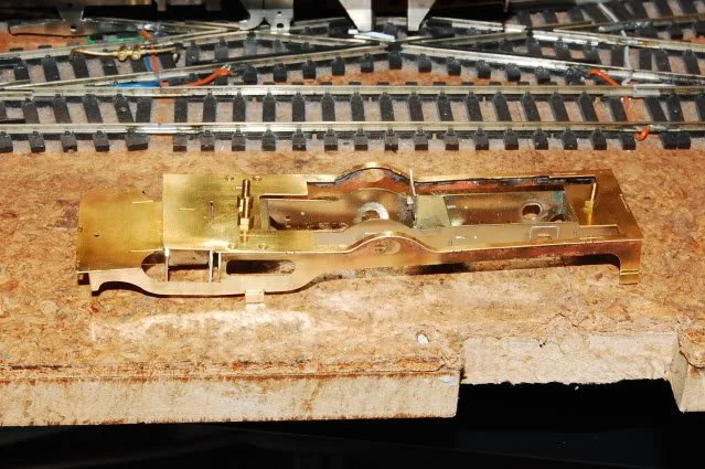 Close-up of a brass or metal model train chassis on a workbench with model train tracks in the background.