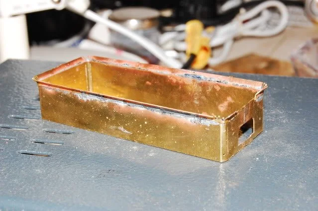 A brass or gold-colored rectangular metal dustpan with a handle opening, resting on a workbench or table surface.