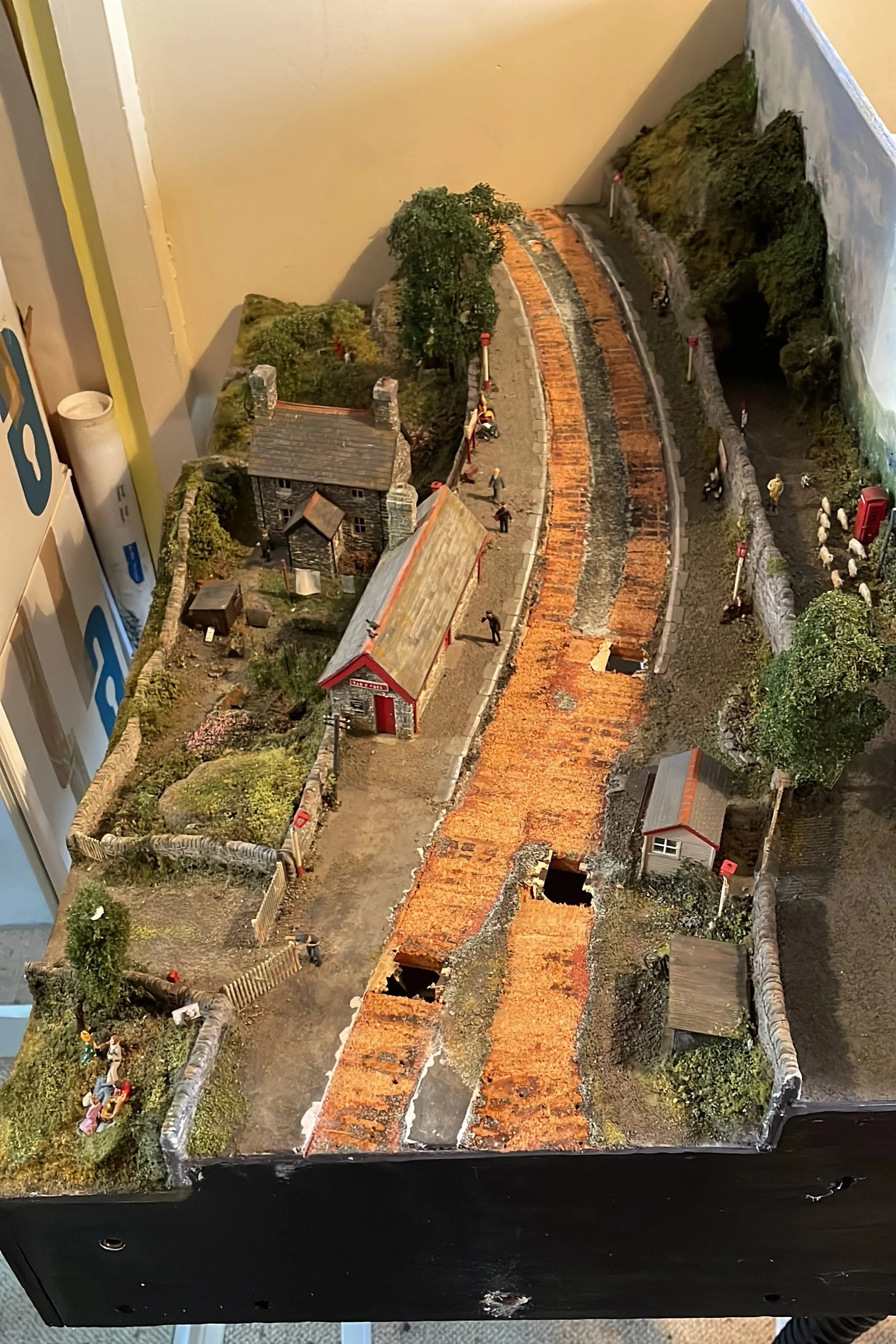 Detailed miniature model of a rural train station scene with buildings, trees, people, and a railway track, some of which is under construction or repair.