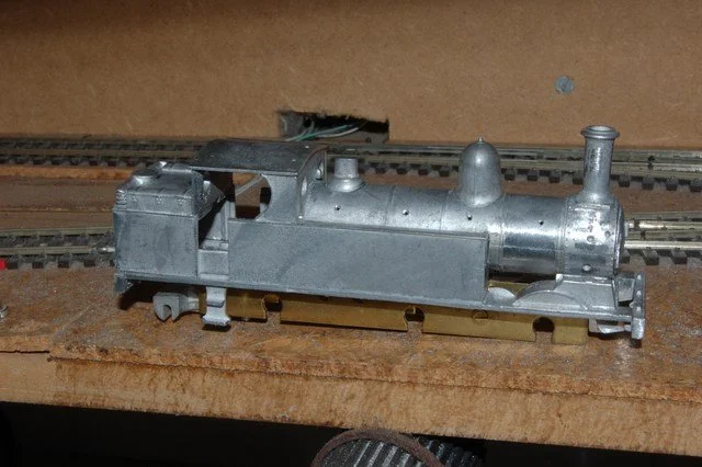 A metal model of a steam locomotive on a wooden surface with train tracks in the background.
