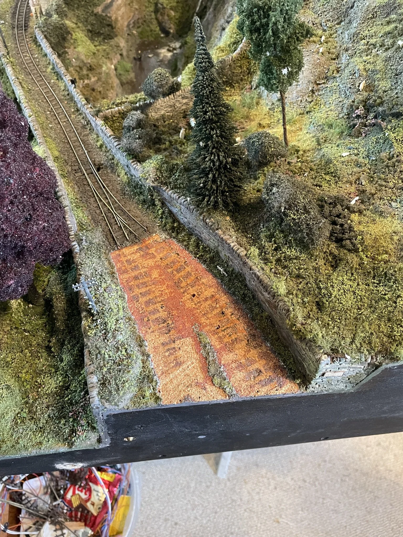 A detailed miniature landscape scene with railway tracks, trees, rocks, and a brick road, creating a realistic outdoor setting.
