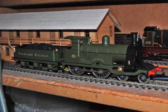 Model green Highland Railway steam locomotive on a model train track with a wooden train station in the background.