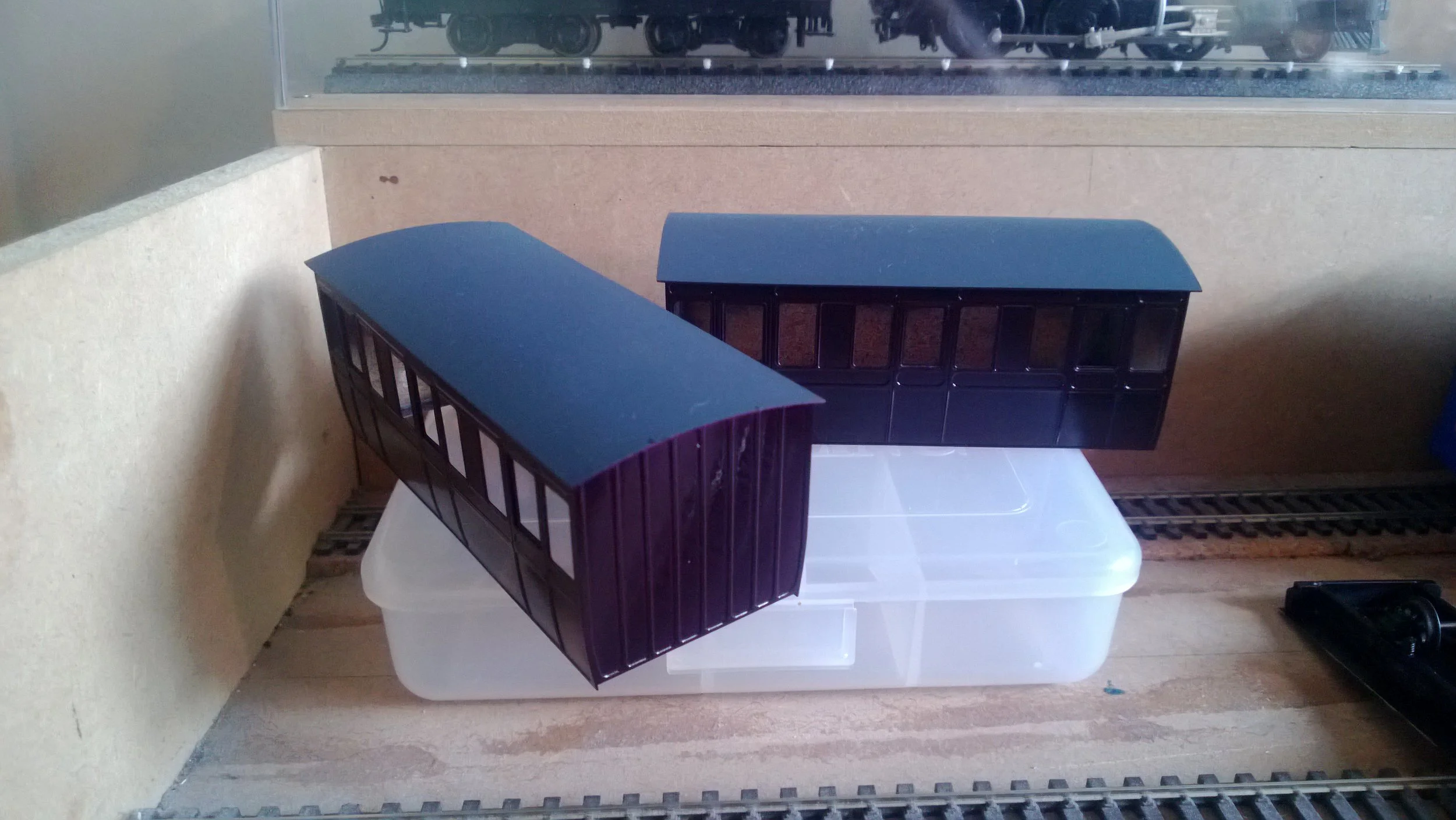 Two model train cars, one purple and one black, placed on a plastic container with a model train track in the background.