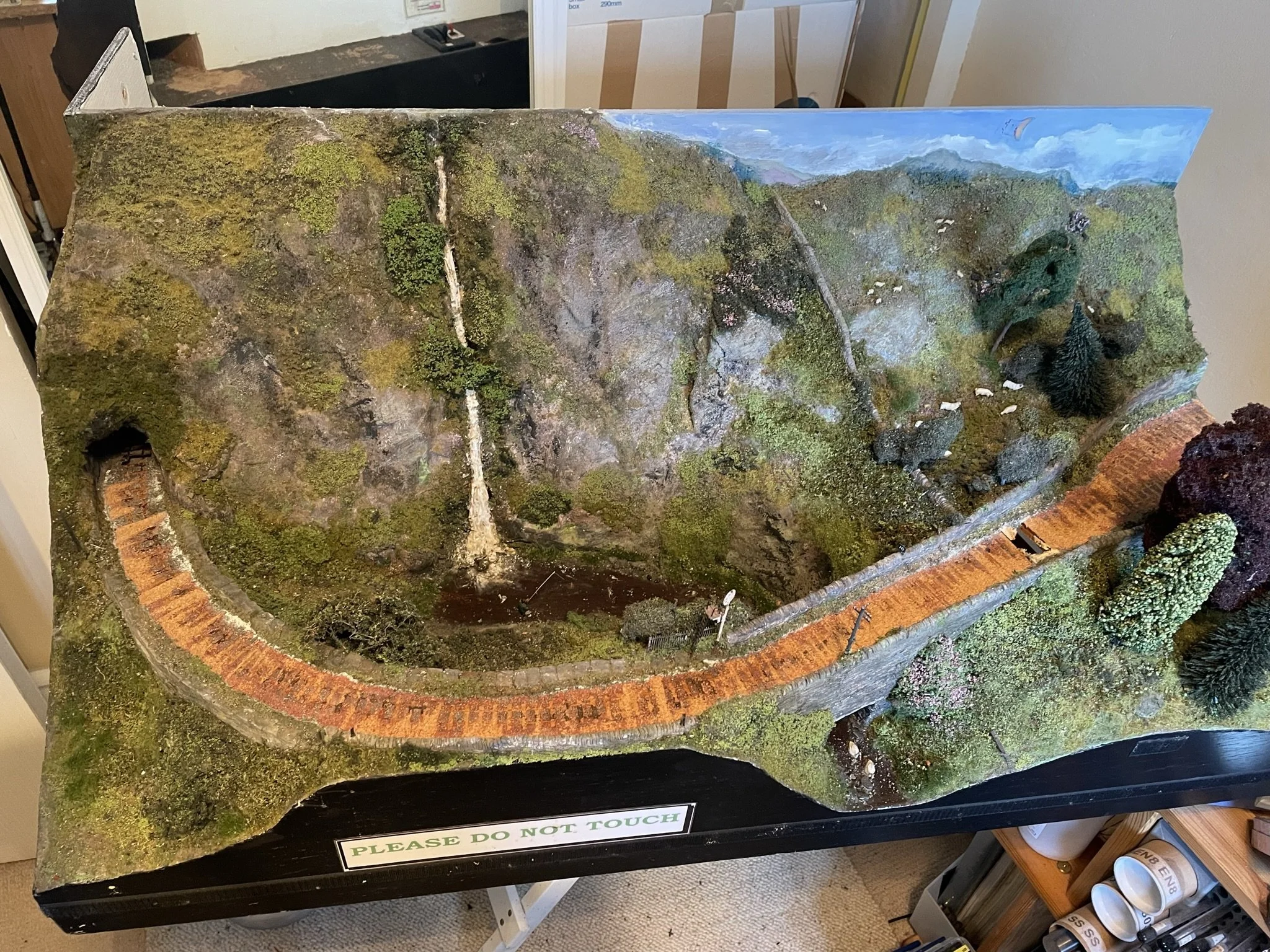 A detailed miniature model of a natural landscape with trees, rocks, a water cascade, and a brick pathway in a diorama style, with a sign saying "Please Do Not Touch".