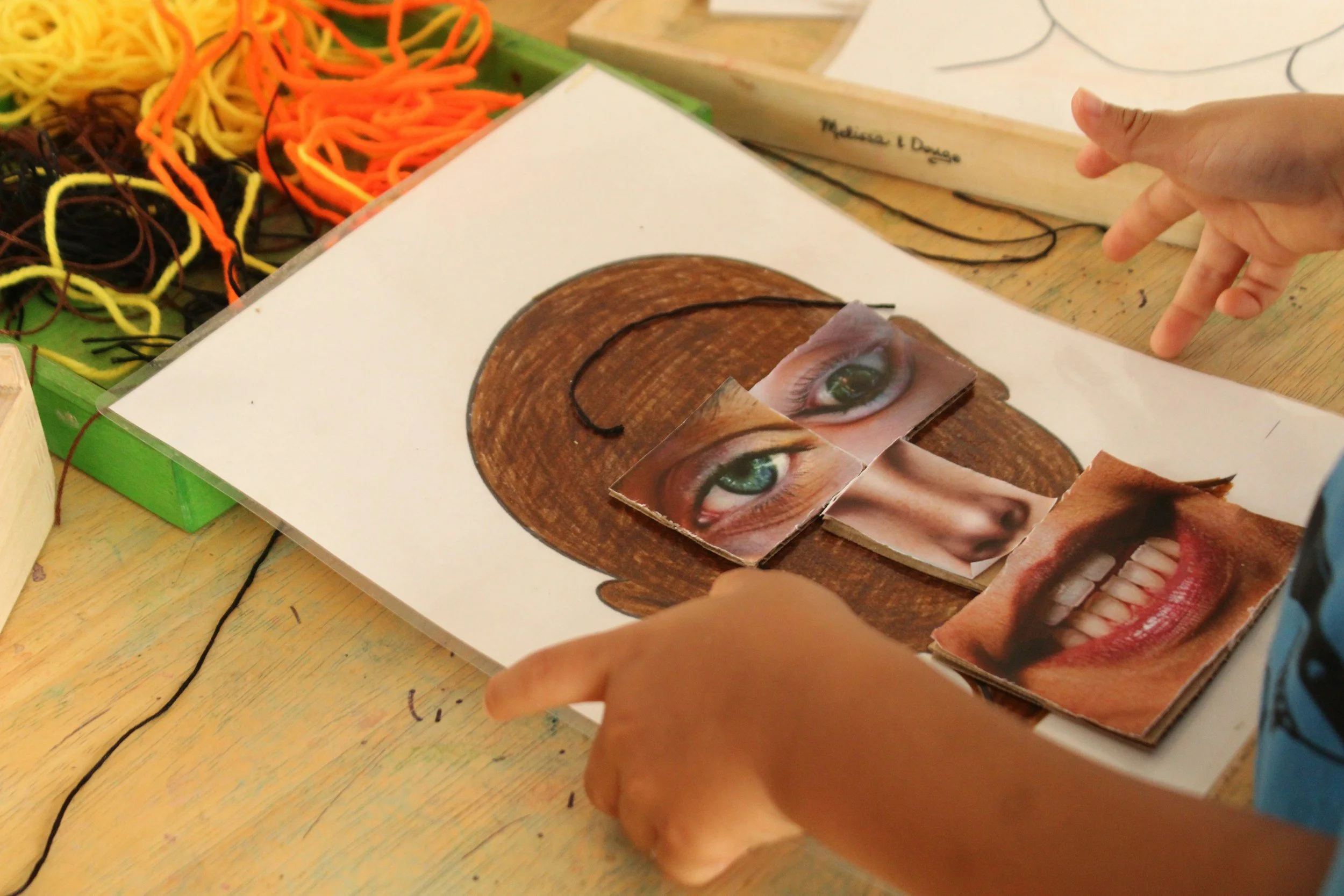Children are working on a collage project involving eyes and smiles, with various pictures and paper on a wooden desk.