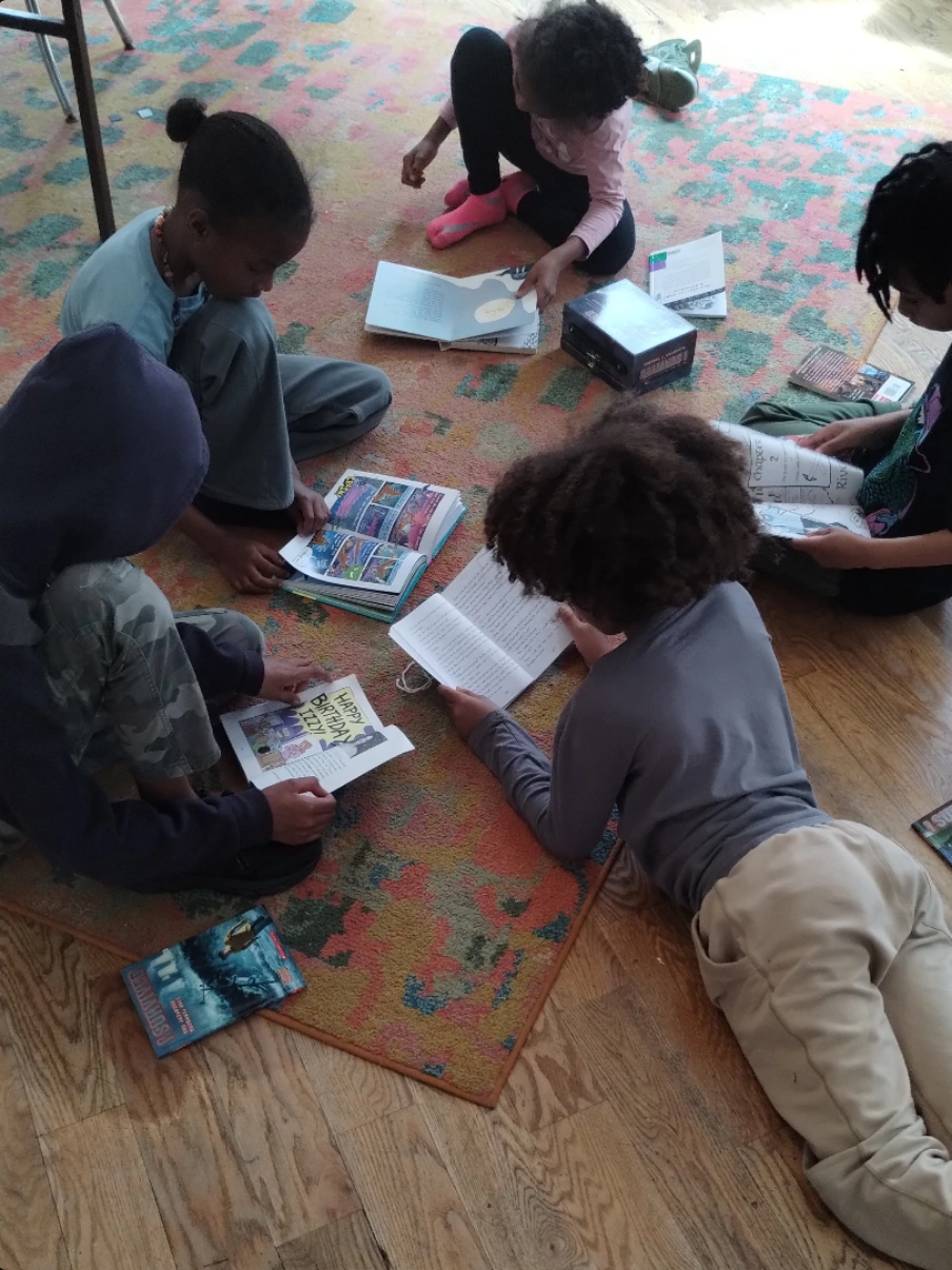 Five children sitting and lying on a colorful rug, reading books. The children are engaged with their books, which include pictures and text, in a room with wooden flooring.