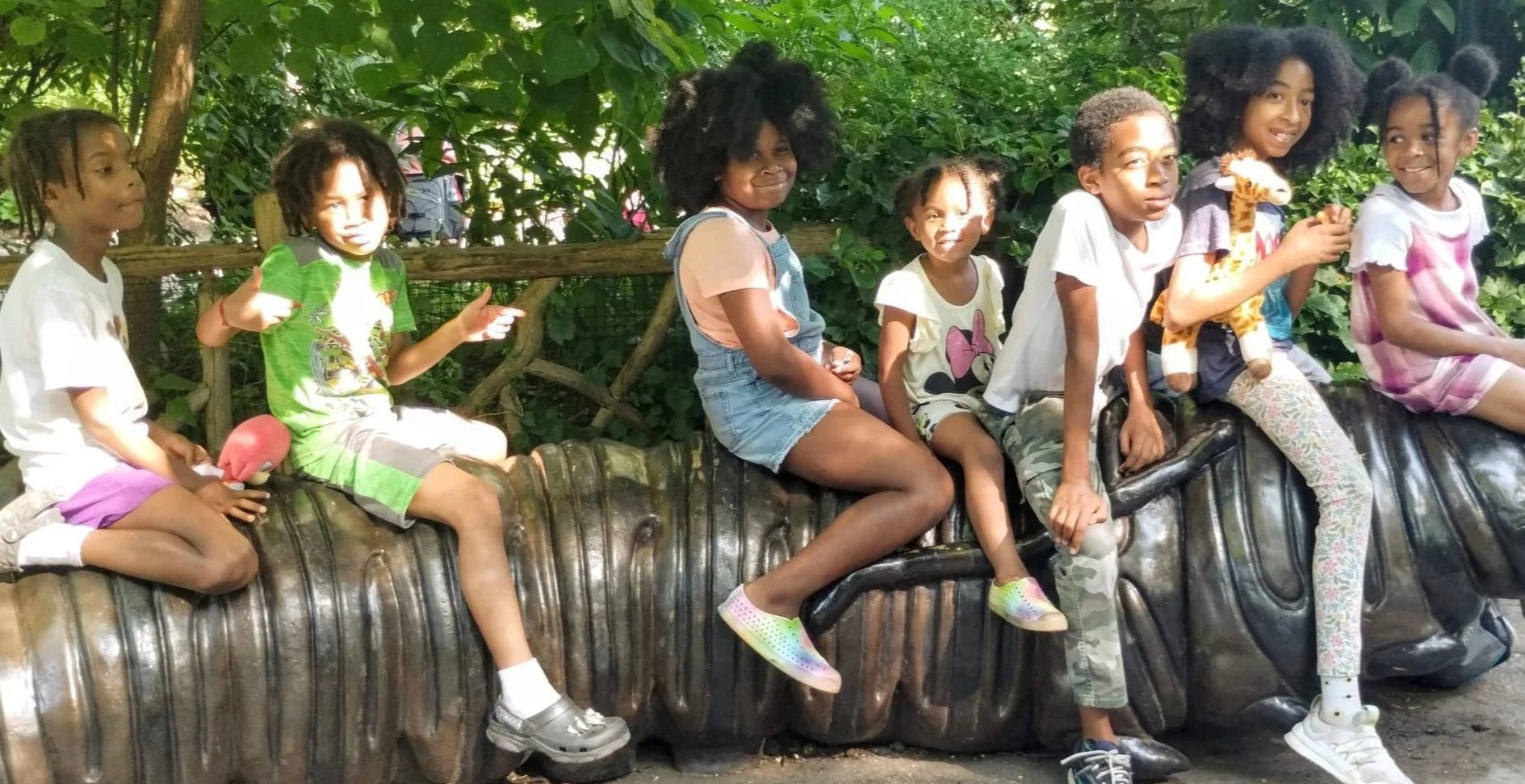 Group of children sitting and playing on a large statue of a hippopotamus outdoors surrounded by green foliage.