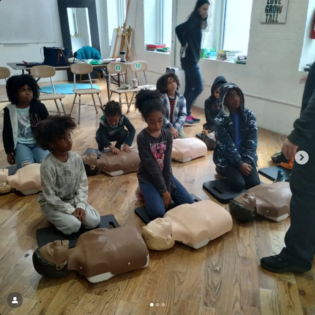 Children participating in a CPR training class, practicing on mannequins on a wooden floor, with a trainer giving instructions.
