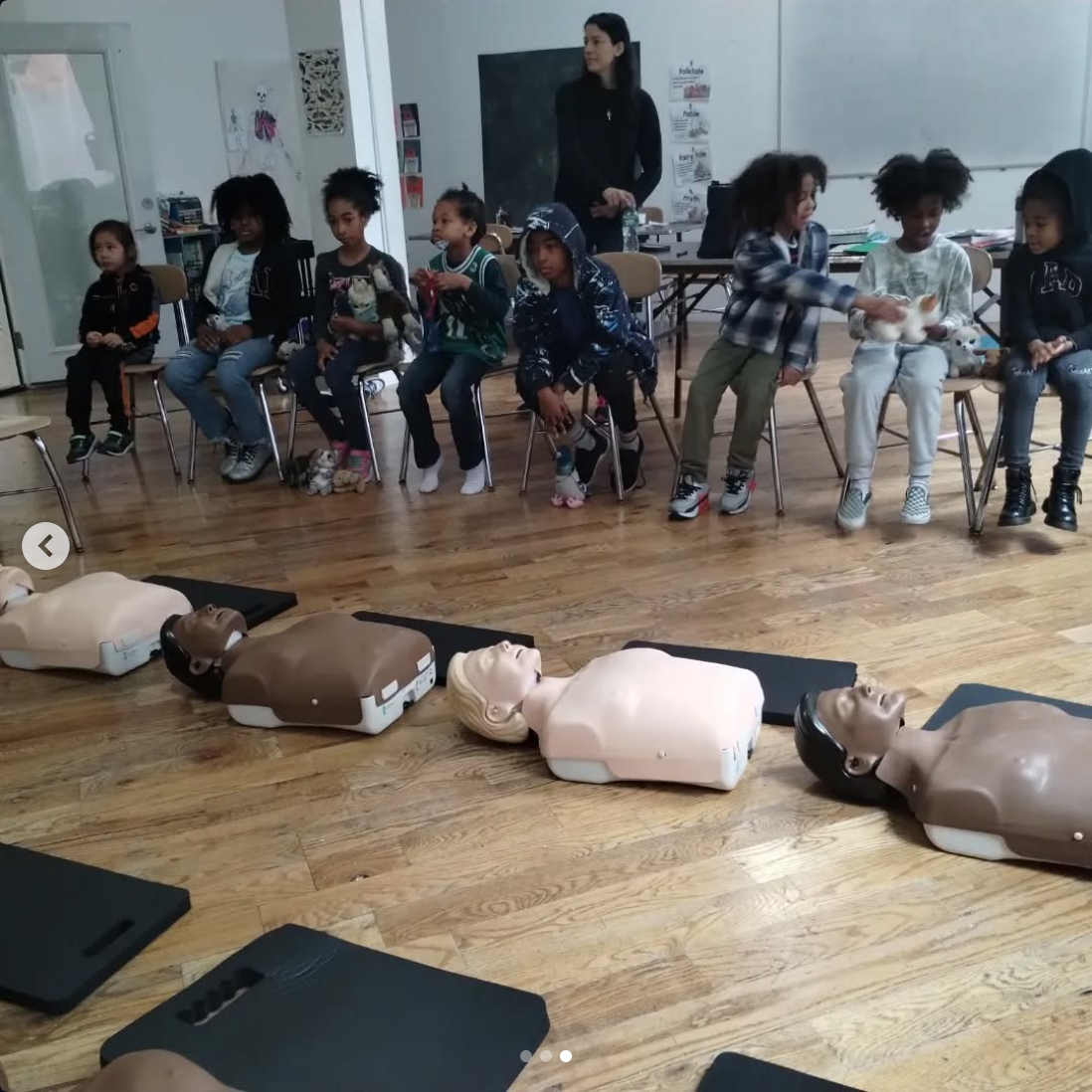 A group of children sitting in a classroom, observing CPR mannequins on the floor for a first aid training session, with a woman standing nearby.
