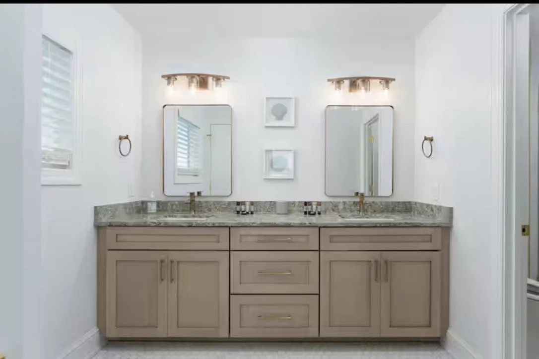 Thoughtful bathroom material selections bring warmth and sophistication to this Kure Beach coastal home, blending soft natural tones, brass details, stone surfaces, and timeless tile for a spa-like feel.