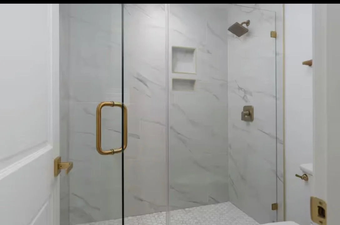 Walk-in shower design with marble-look tile and brass hardware in Kure Beach beach house.