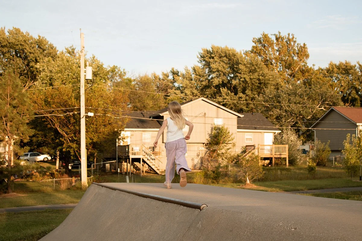 A young girl with long hair, wearing a white top and purple pants, running on a skate park ramp during sunset, with trees and houses in the background.