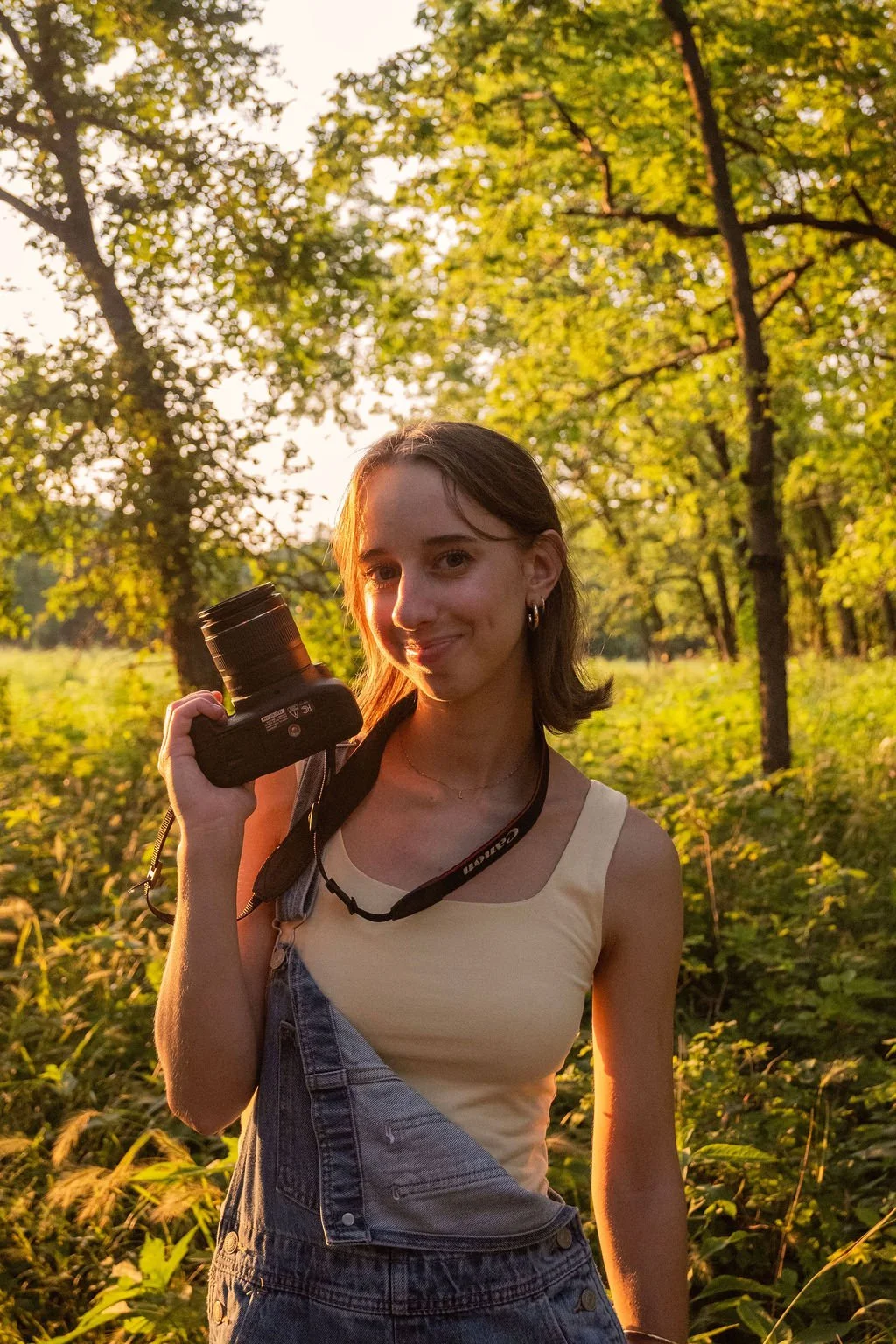 Young woman holding a camera outdoors in a lush, green forest during sunset.