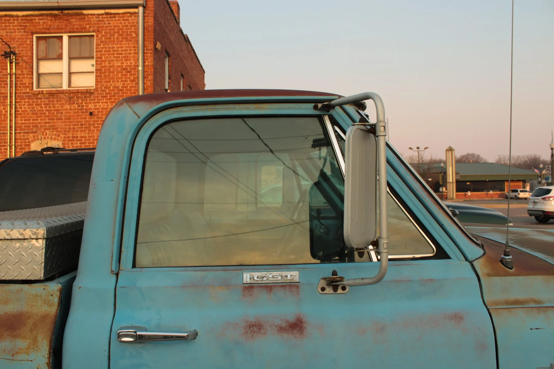 Close-up of a vintage light blue pickup truck with rust spots, metal side mirror, and view through the windshield showing overhead power lines and a parking lot with other cars, brick building, and a clear sky in the background.
