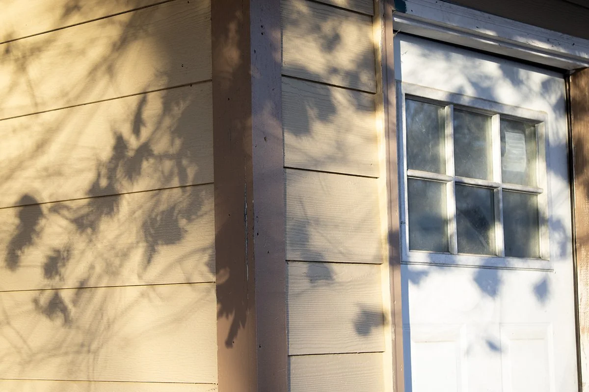 A corner of a house with horizontal beige wooden siding and a white garage door with four small window panes. Shadows of tree branches are cast on the siding.