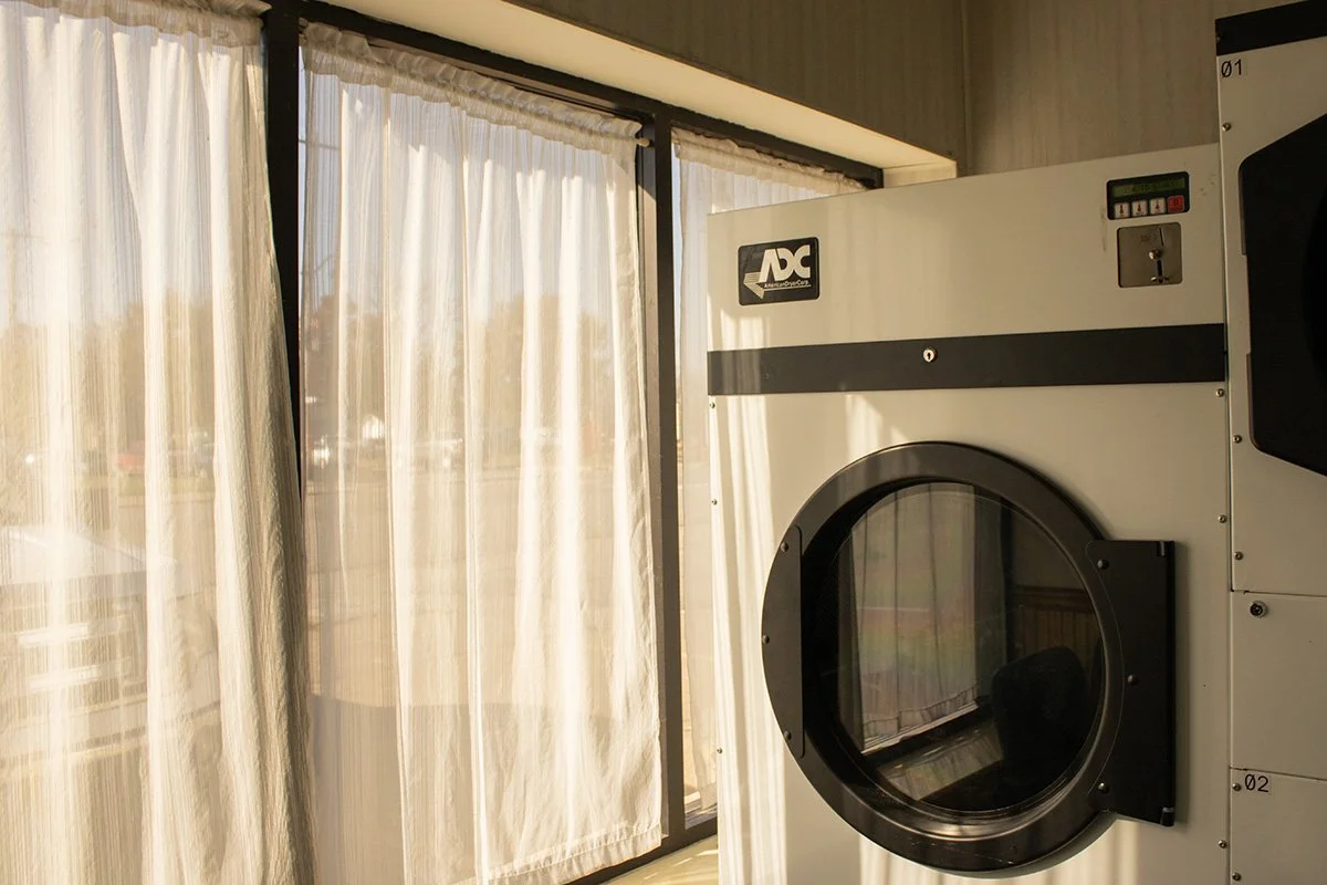 An industrial laundry dryer with a circular door, located near a large window with white curtains, allowing sunlight to enter the room.