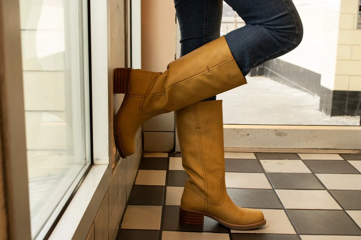 A person wearing yellow tall leather boots and dark jeans standing in front of a window and door.