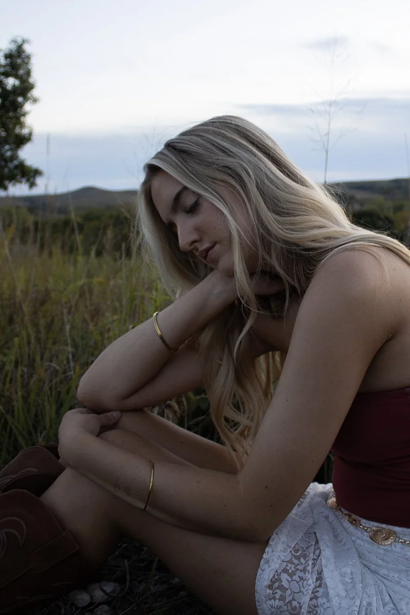 A woman with long blonde hair sitting outdoors in a field with mountains in the background, eyes closed, head resting on her knee, wearing a maroon top and white lace skirt.