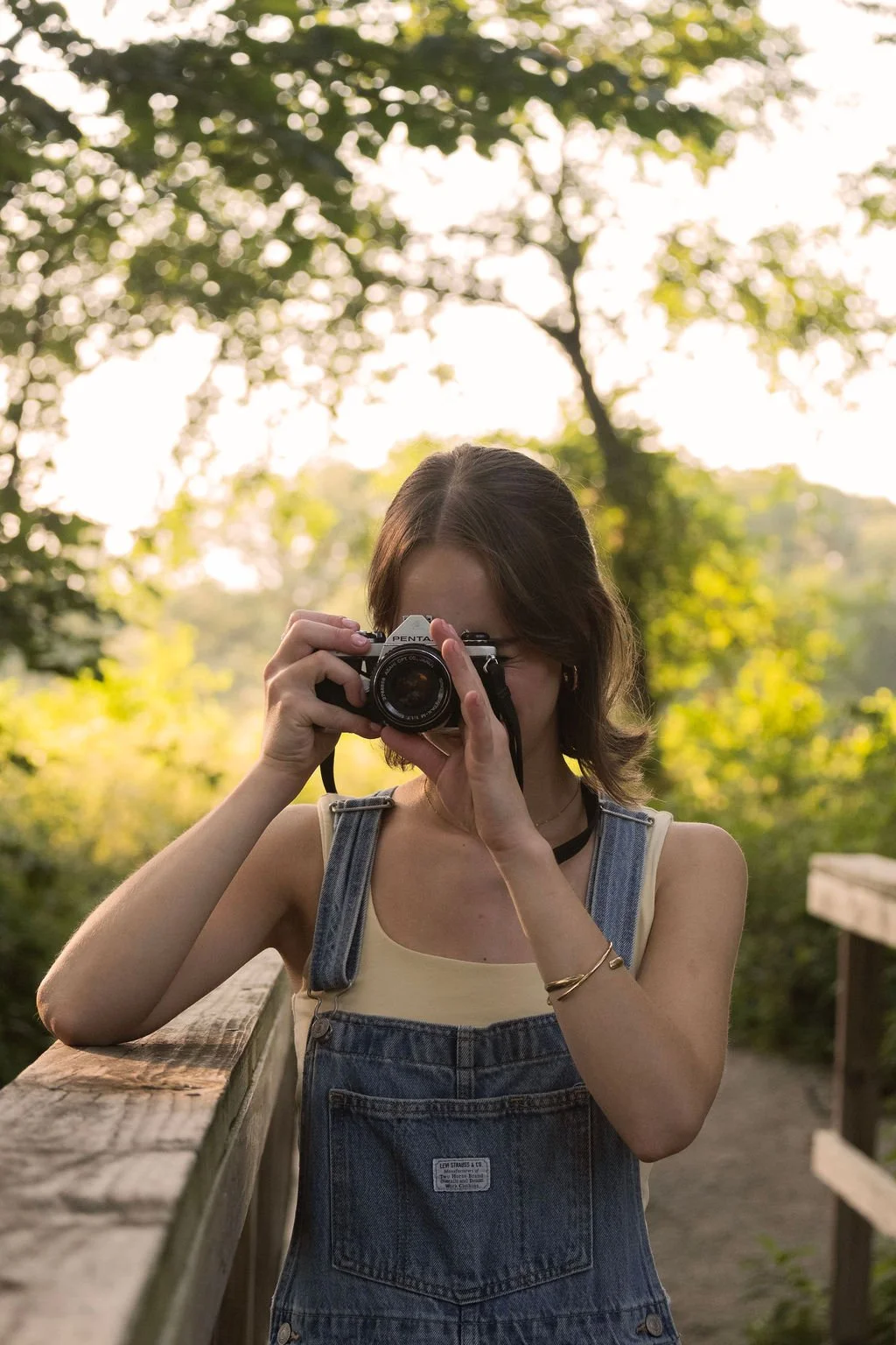 A woman taking a photo with a camera outdoors during daylight, surrounded by green trees and foliage.
