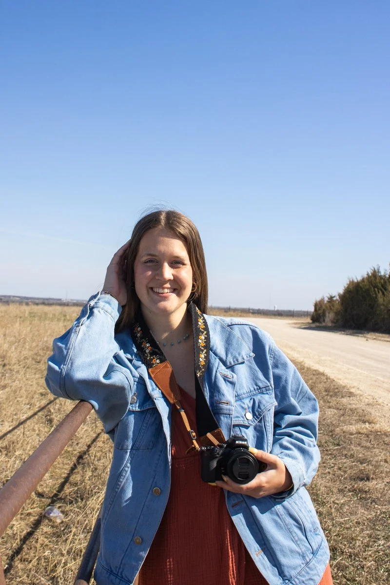 A young woman with brown hair smiling and posing outdoors, holding a camera, wearing a denim jacket over a rust-colored dress, on a sunny day in a rural area with a dirt road and open field.