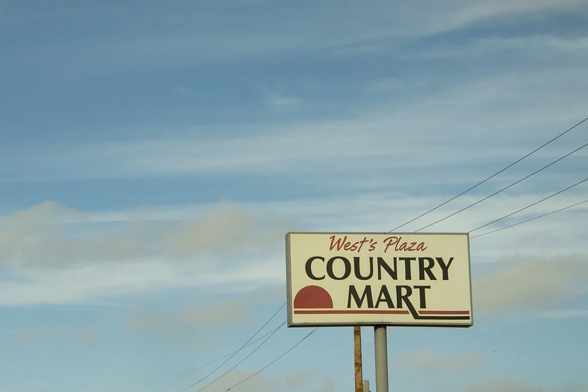 A sign for West's Plaza Country Mart against a partly cloudy sky.