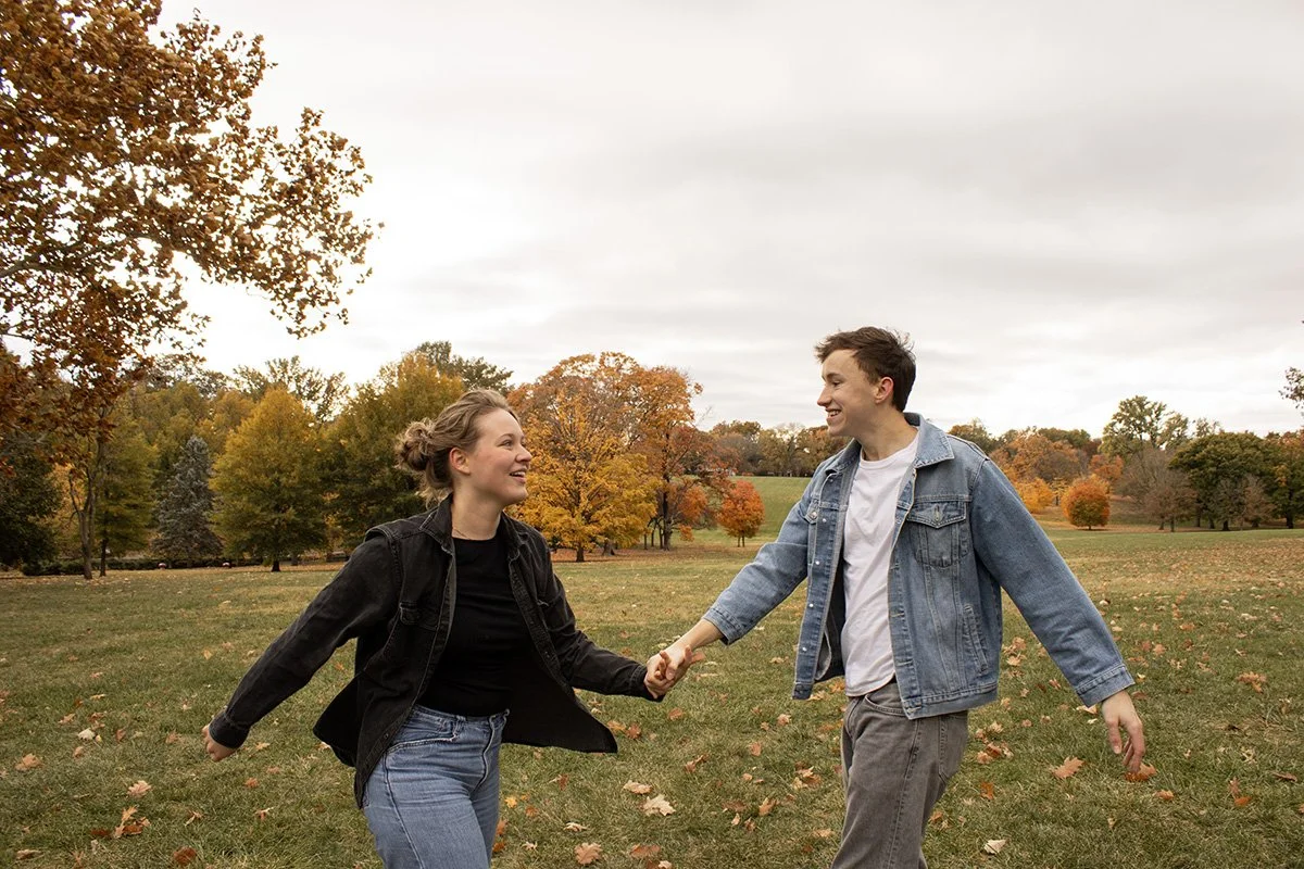 A young couple is holding hands and smiling at each other in a park during fall, with trees showcasing colorful autumn leaves in the background.