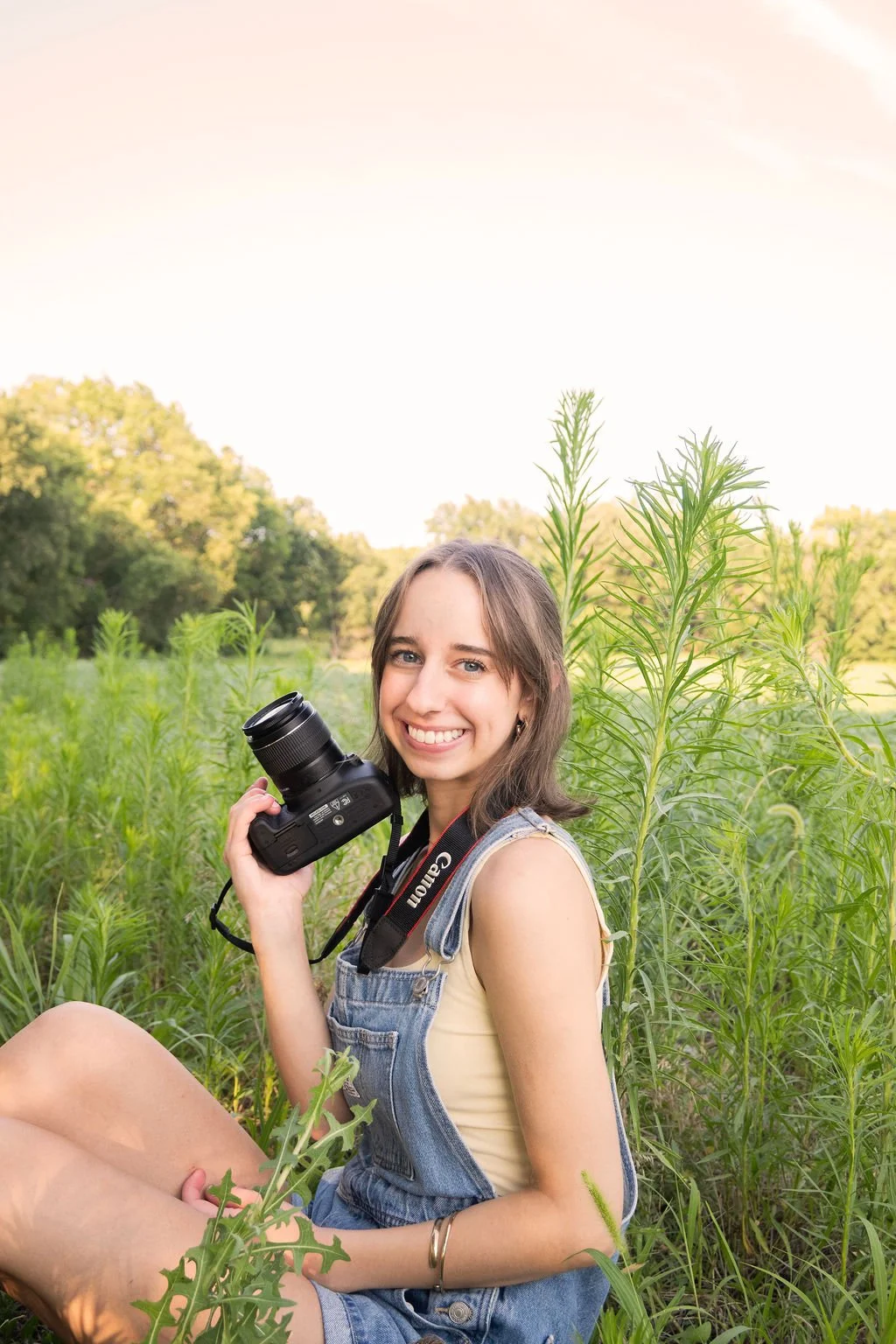 A young woman sitting in a field of tall green plants, smiling, holding a DSLR camera, wearing denim overalls and a yellow tank top with sunlight and trees in the background.