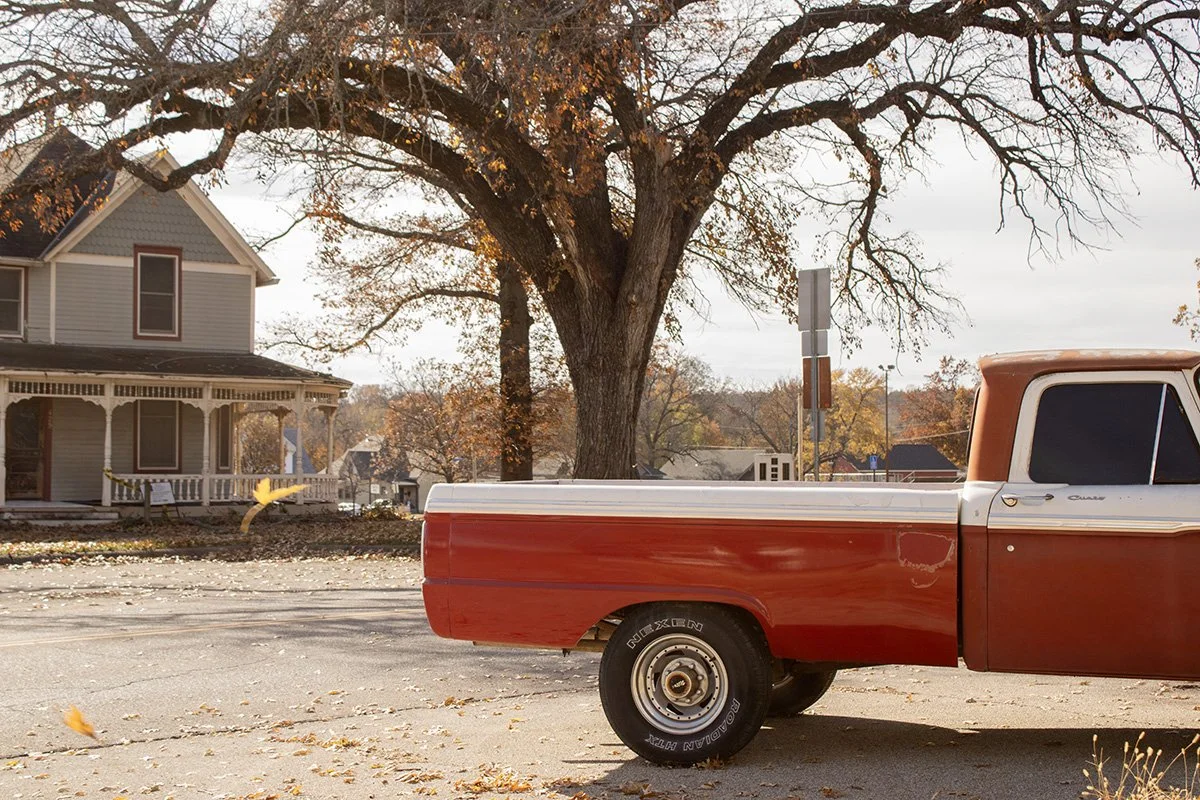 A vintage red pickup truck parked on a street in front of a house with a large tree and autumn leaves.
