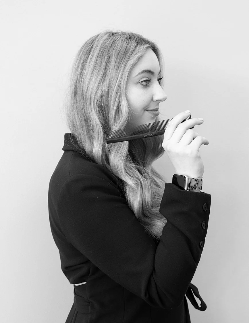 Side view of a woman with long wavy hair holding a comb near her face, wearing a dark blazer and a smartwatch on her left wrist, standing against a plain background.