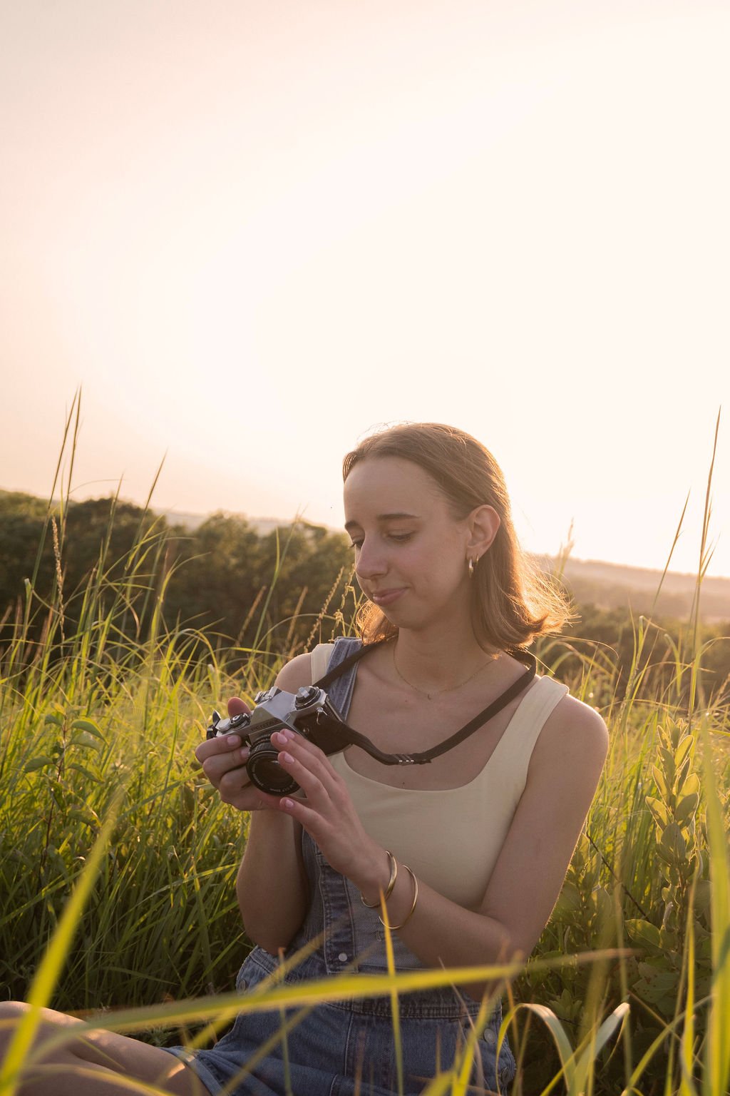 A young woman sitting in tall grass during sunset, holding a camera and looking down at it.