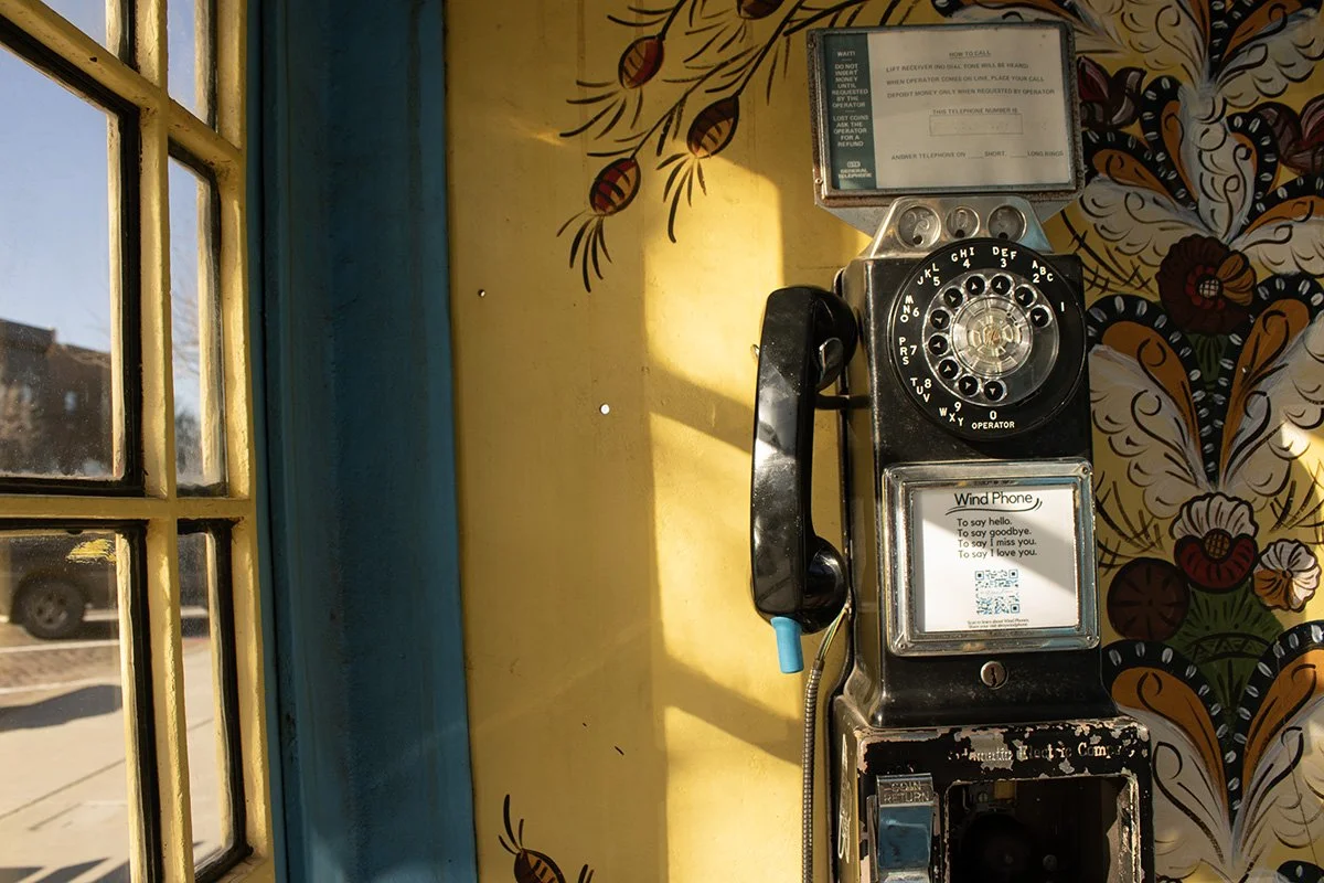 Vintage black rotary payphone mounted on a yellow wall with floral and butterfly decorations, next to a window with yellow framing.